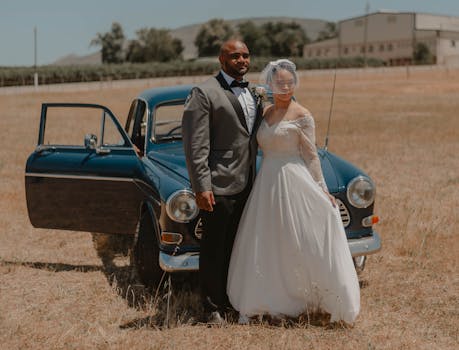 Bride and groom pose elegantly beside a classic vintage car in a serene field.