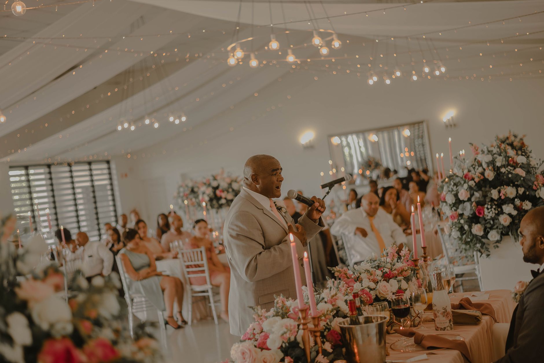 A man gives a speech at a wedding reception surrounded by guests and floral arrangements.