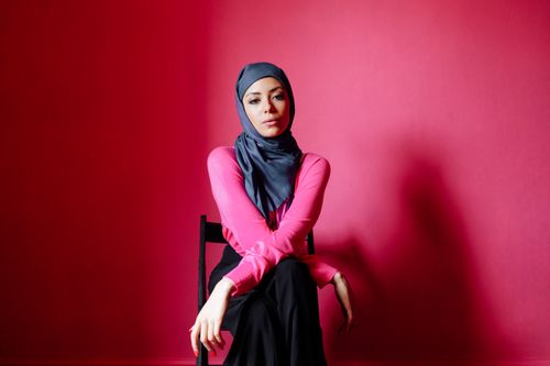 Free A woman in a hijab sits gracefully against a vibrant red background, exuding confidence and style. Stock Photo