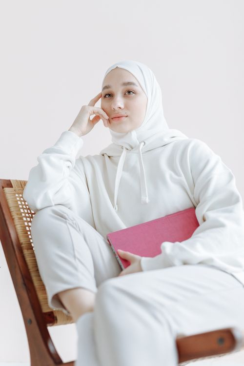 Free A woman in a white hijab and hoodie sits thoughtfully holding a pink book. Stock Photo