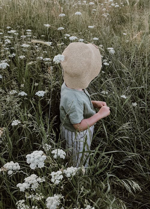 Side view of unrecognizable kid in casual clothes and straw hat standing on meadow with white flowers while looking away in summer