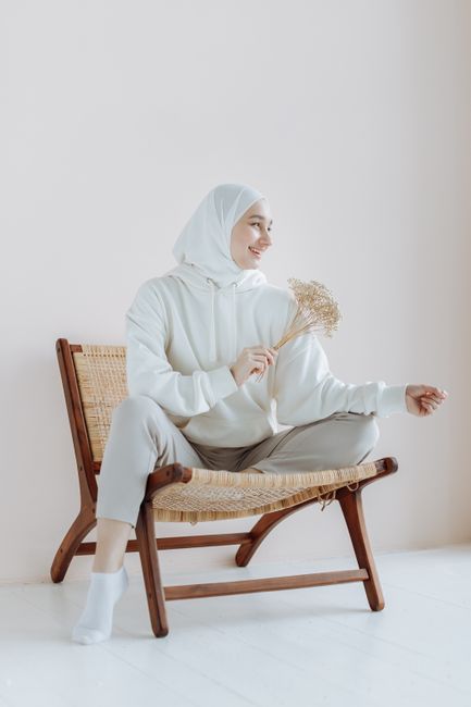 A woman in a hijab smiles while seated on a wooden chair, holding dried flowers in a minimalistic setting.