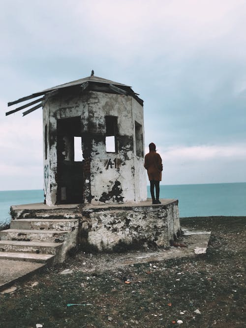 Back view full length of unrecognizable female in outerwear standing near abandoned shabby alcove and admiring sea