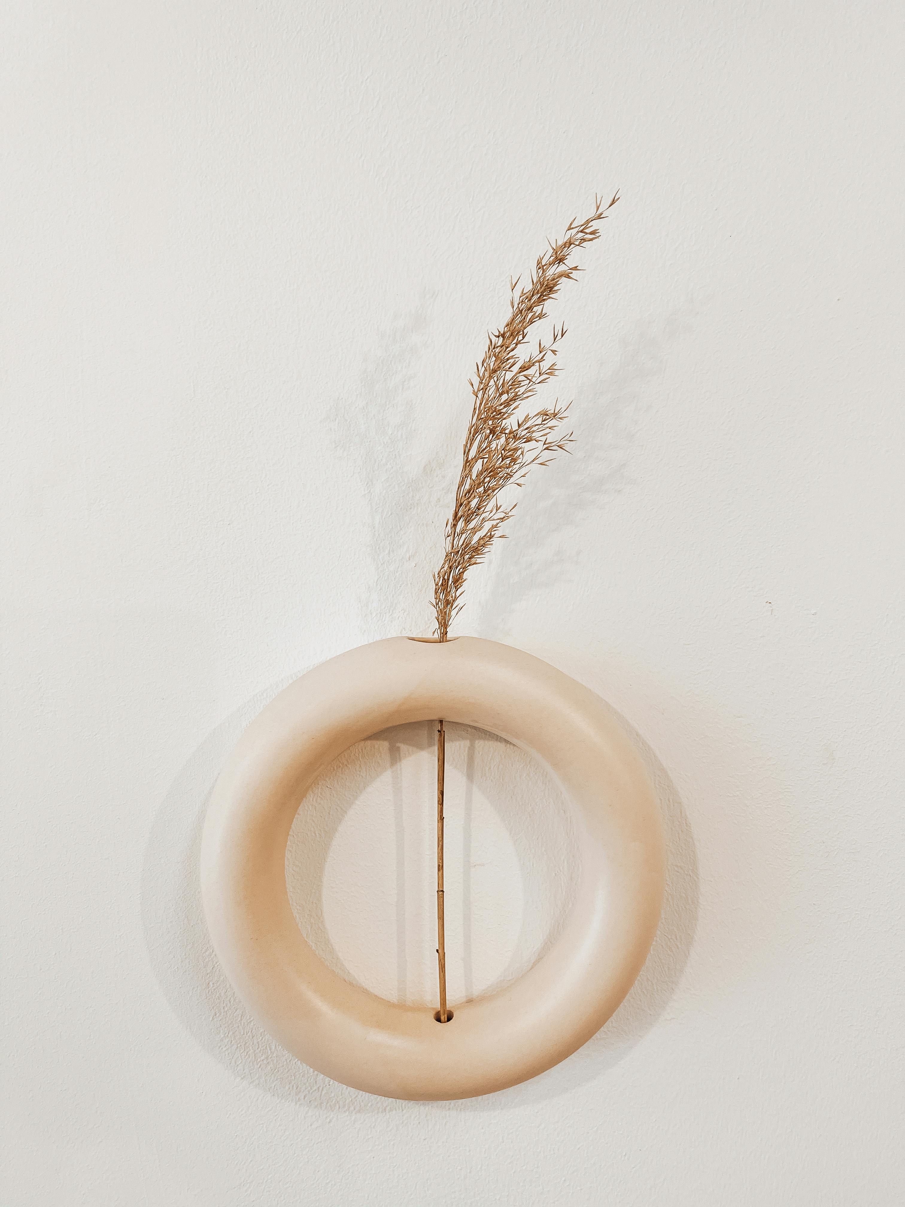 A beige circular ceramic vase with a dried plant on a plain white background, exhibiting minimalist design.