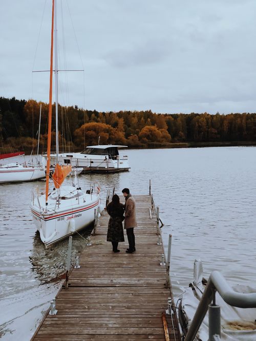 Free A couple stands on a wooden dock by a lake with sailboats, capturing the beauty of autumn in Belarus. Stock Photo