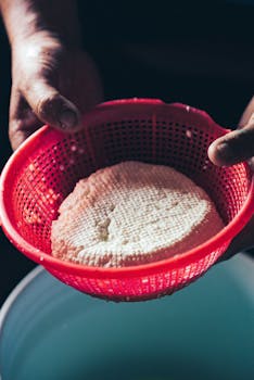 High angle of crop anonymous person standing with homemade cheese in colander above bucket