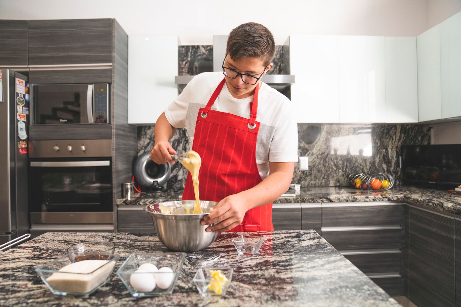 Teenager in a modern kitchen wearing a red apron while whisking ingredients in a stainless steel bowl.
