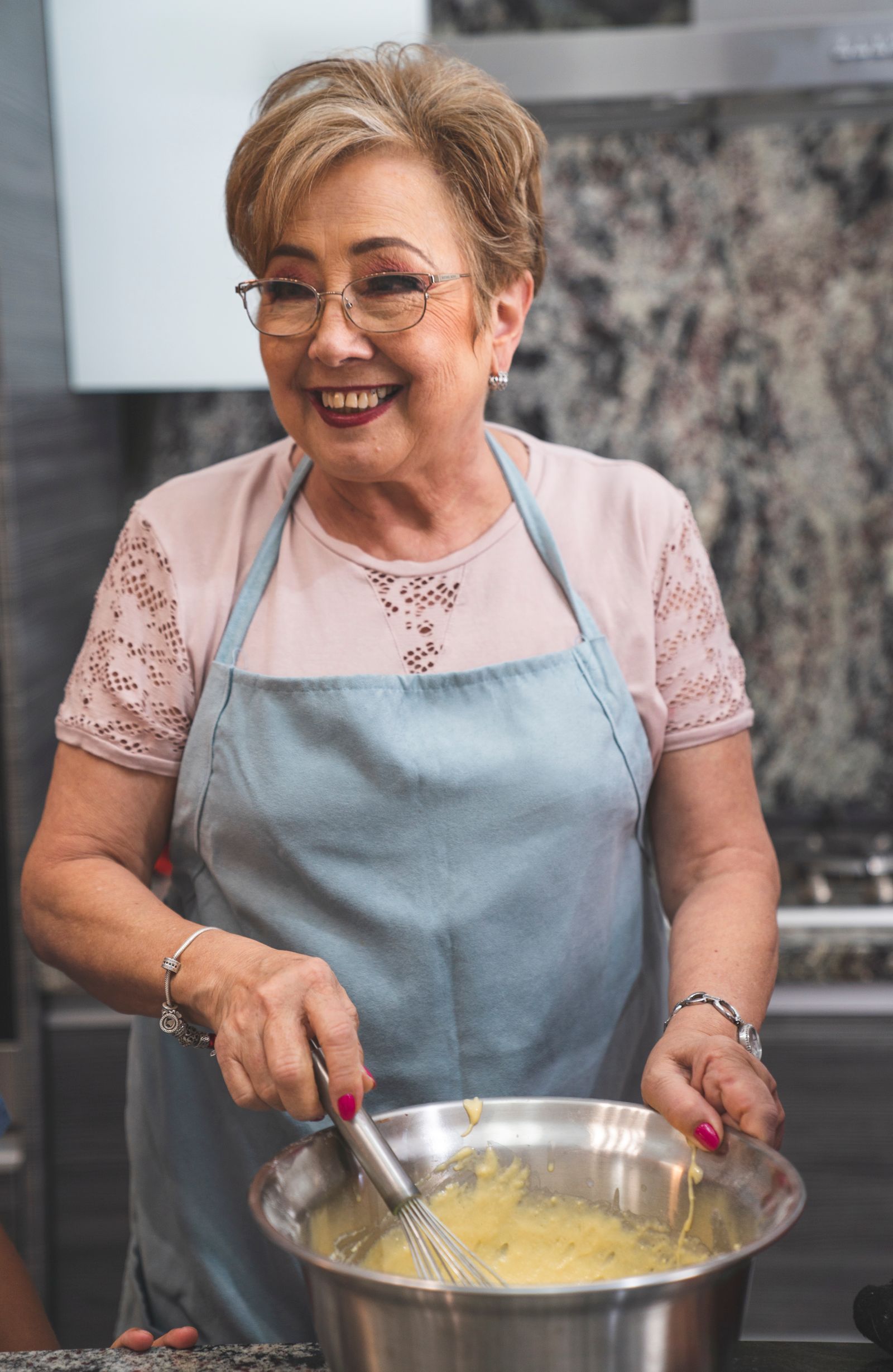 A smiling older woman with glasses and a blue apron whisking batter in a steel bowl