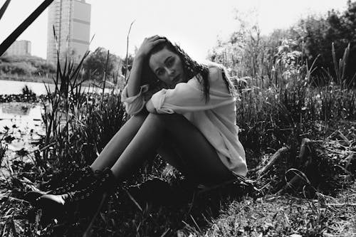 Black and white full body of gorgeous female in casual clothes sitting on grassy coast near river in park while touching hair and looking at camera