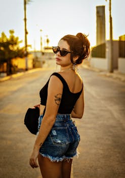 Woman with sunglasses and tattoos in a stylish pose during sunset on an urban street.