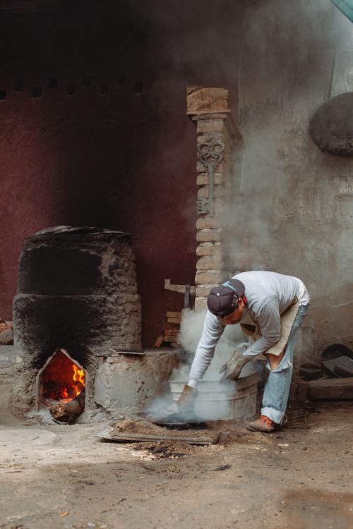 Free A man working in a rustic environment with fire and smoke. Wearing gloves and long sleeves, he tends to a metal drum. Stock Photo