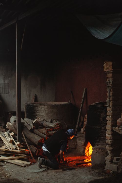Free A man working in a rustic workshop, tending to a brick fireplace surrounded by firewood. Stock Photo