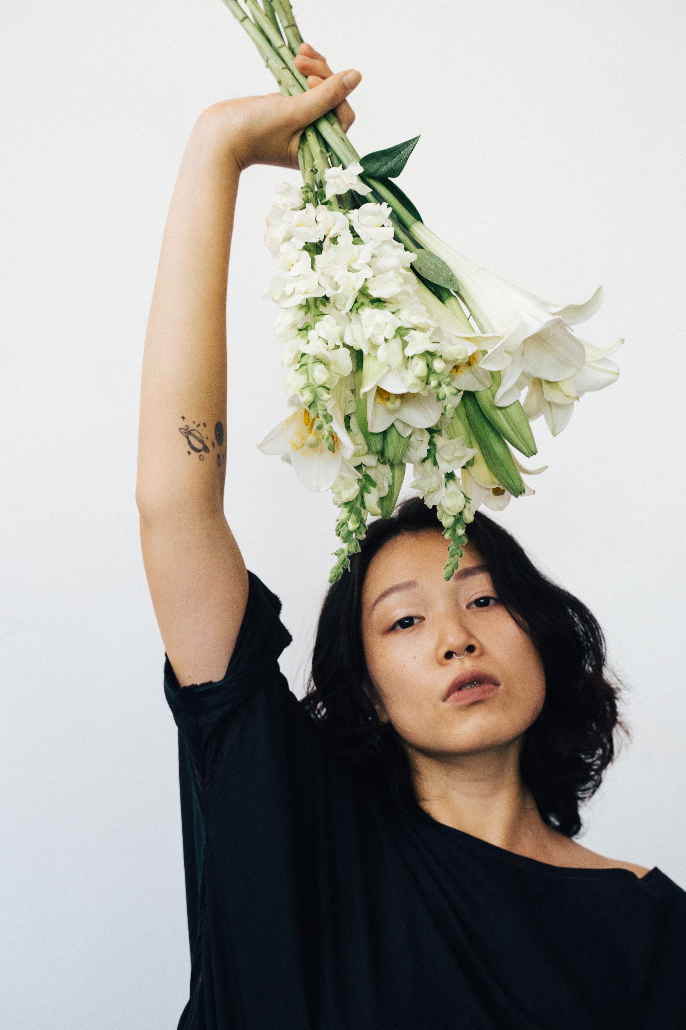 Asian woman in black shirt holding white flowers, arm raised elegantly.