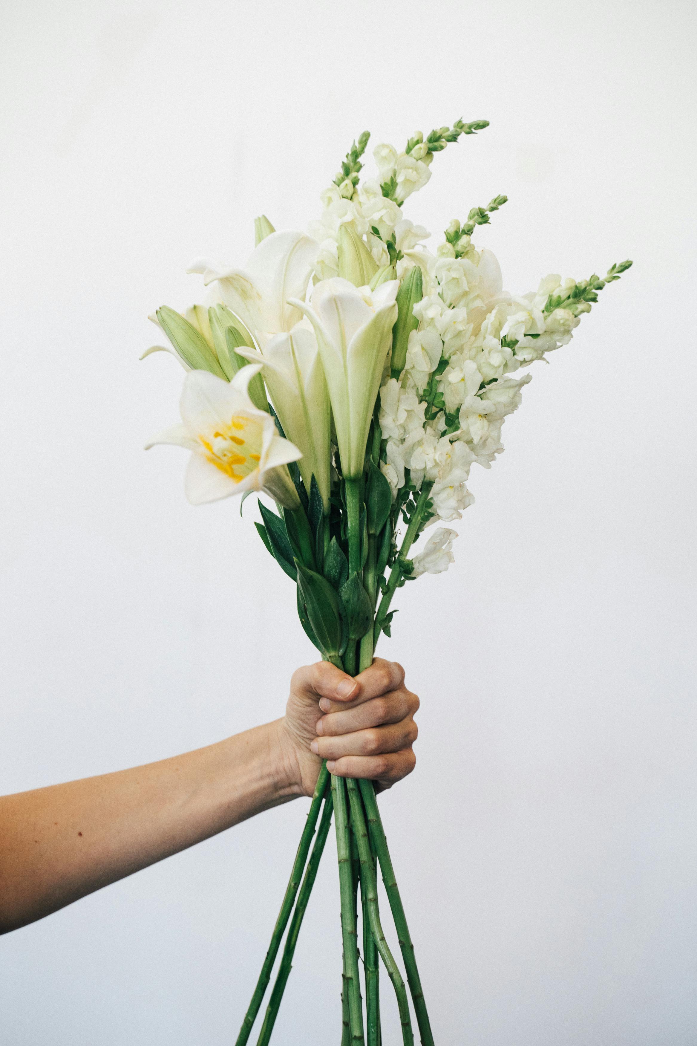 A hand holding a delicate bunch of white lilies and snapdragons against a plain background.