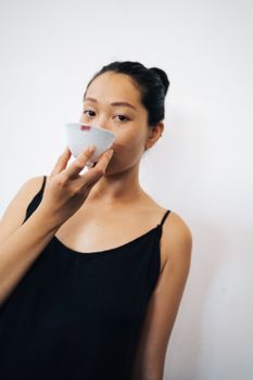 A young woman in a black dress enjoying a drink from a ceramic cup indoors against a neutral background.
