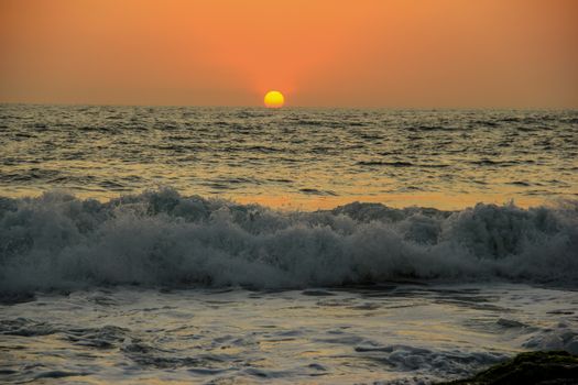 Experience a breathtaking Bali sunset with waves crashing on the shore under an orange sky.