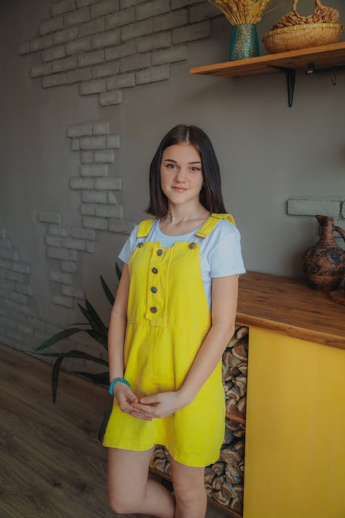 Gentle teenager in yellow overall dress looking at camera against table with vase at home