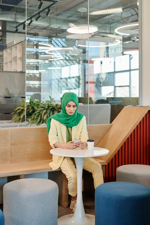 Free Young woman in a hijab using her smartphone at a modern indoor café setting. Stock Photo