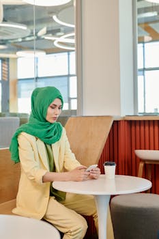Woman in hijab using smartphone in a modern cafe setting, enjoying a coffee break.