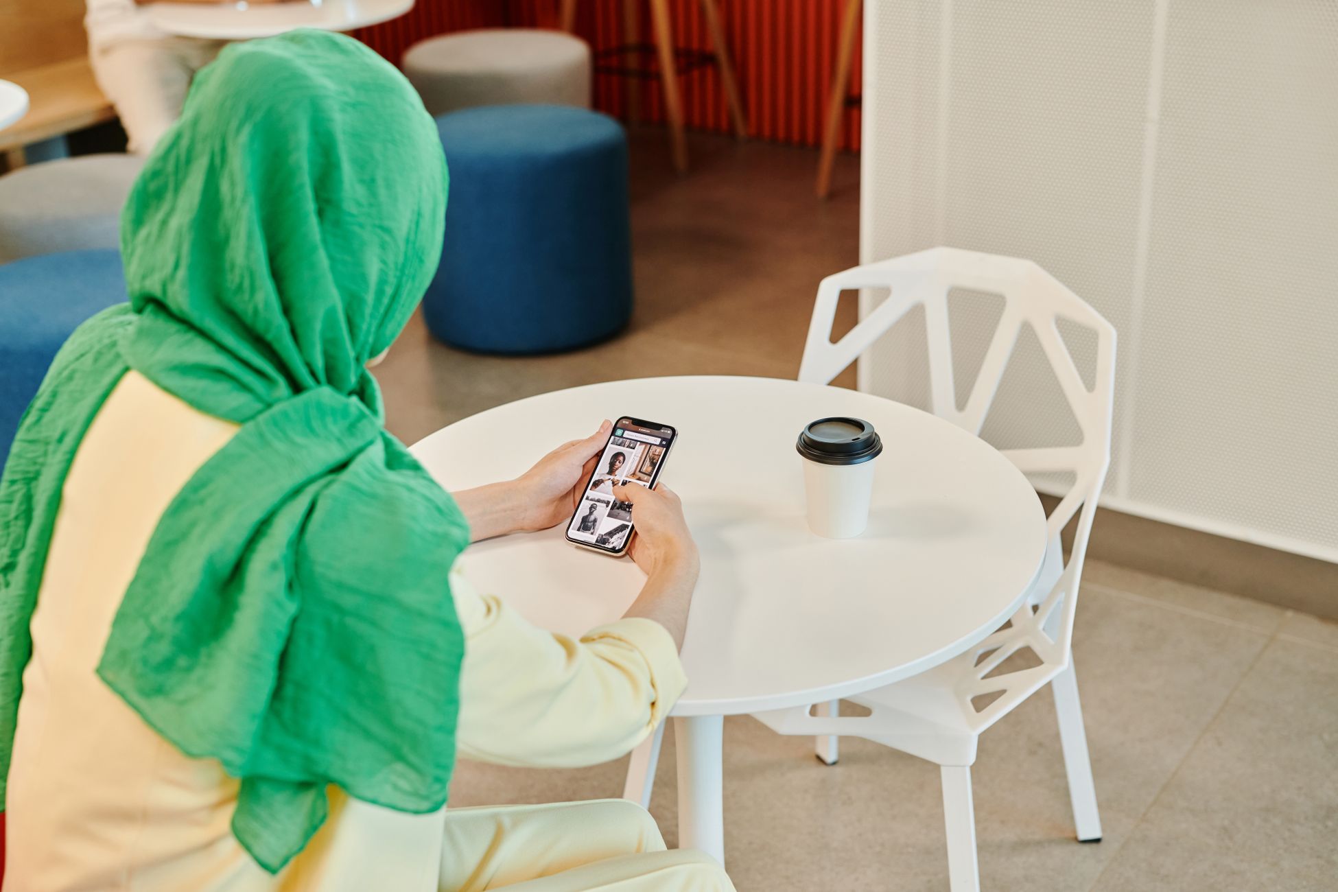A woman wearing a green hijab is scrolling on her smartphone while sitting at a cafe table.