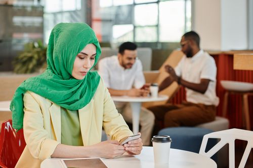 Free Young woman in a green hijab using smartphone at a cafe with colleagues nearby. Stock Photo
