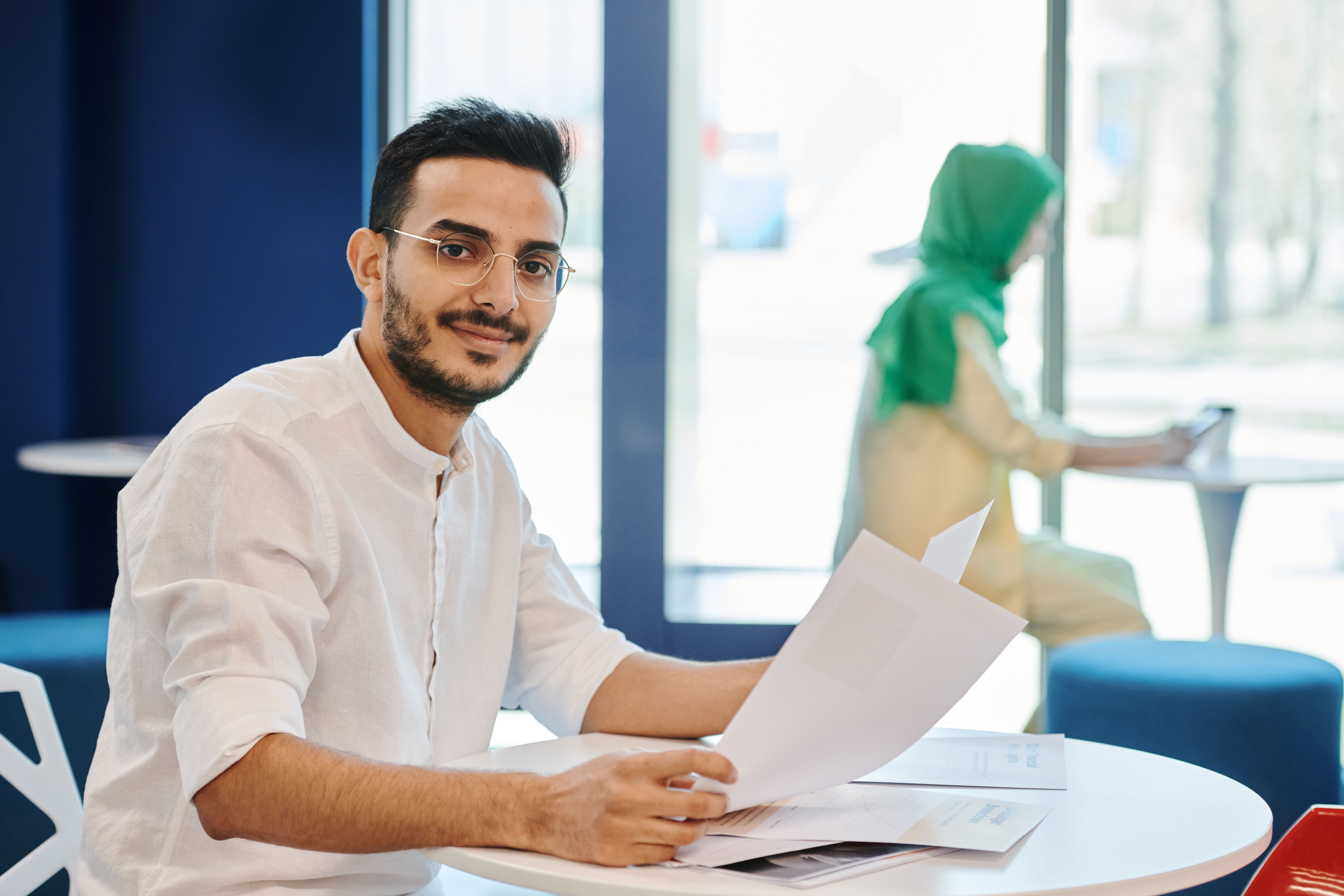 Man with glasses and beard smiling while reviewing documents at table in a modern office setting.