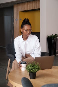 Young businesswoman using a laptop and drinking coffee in a stylish office setting.