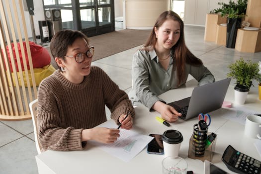 Two women engaged in a casual office meeting, working on laptops and documents.
