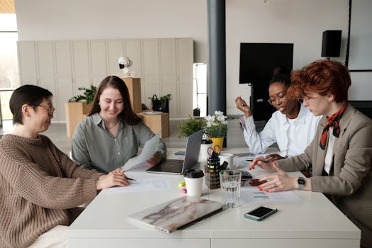 Four diverse women engaged in a collaborative work meeting in a bright, modern office space.