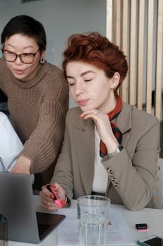 Two women engage in a professional discussion using a laptop in an office setting.