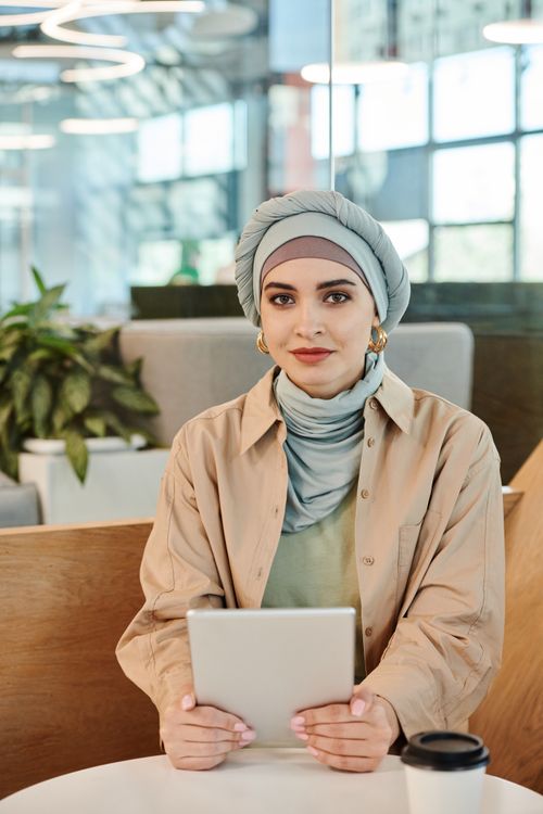 Free Stylish woman in hijab holding tablet at a contemporary cafe table with a cup of coffee and plants. Stock Photo