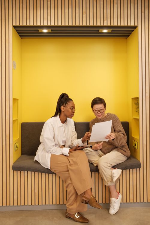 Free Two women engaged in a business discussion, reviewing documents in a modern office nook. Stock Photo