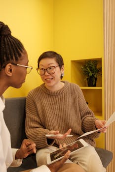 Two women discuss business strategies holding documents and a tablet in a vibrant yellow office.