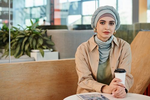 Free Modern woman in hijab at a café holding coffee and tablet, expressing a blend of style and comfort in an indoor setting. Stock Photo
