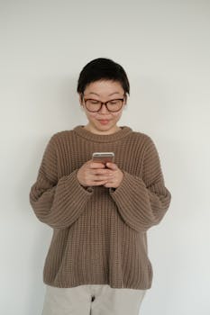 A woman in a brown sweater and glasses intently views her smartphone against a white background.