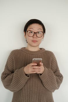 Asian woman with short hair and eyeglasses, standing against white background, wearing a brown sweater and holding a smartphone.