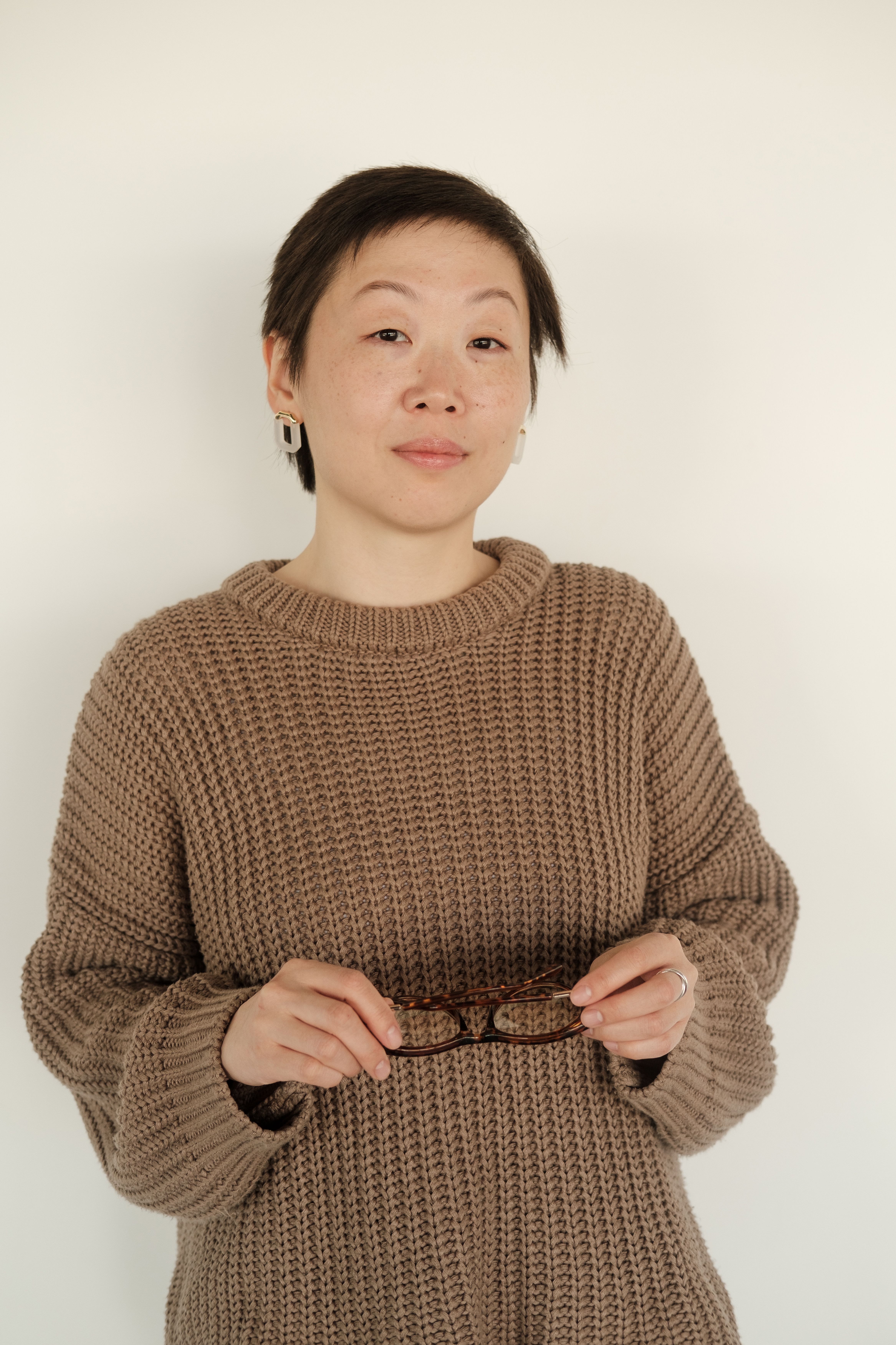 Stylish Asian woman with short hair holding glasses, wearing an elegant knitted sweater.