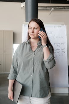 A professional woman holds a laptop while talking on a mobile phone in an office environment.