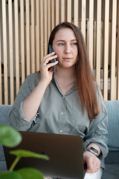 Young woman indoors on a phone call while working on her laptop, showcasing communication and multitasking.