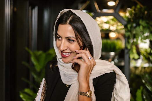 A woman in a hijab smiles while making a phone call in a lush indoor setting.