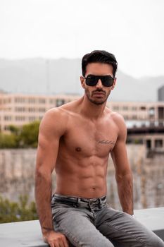 A fit and muscular man sits outdoors, showcasing tattoos and sunglasses.