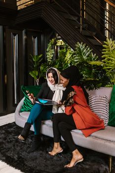 Two women wearing hijabs laughing and looking at a smartphone indoors.