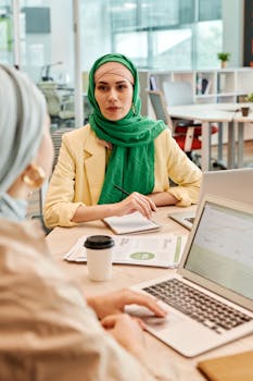 Two women in hijabs discussing business at office table with laptops and papers.