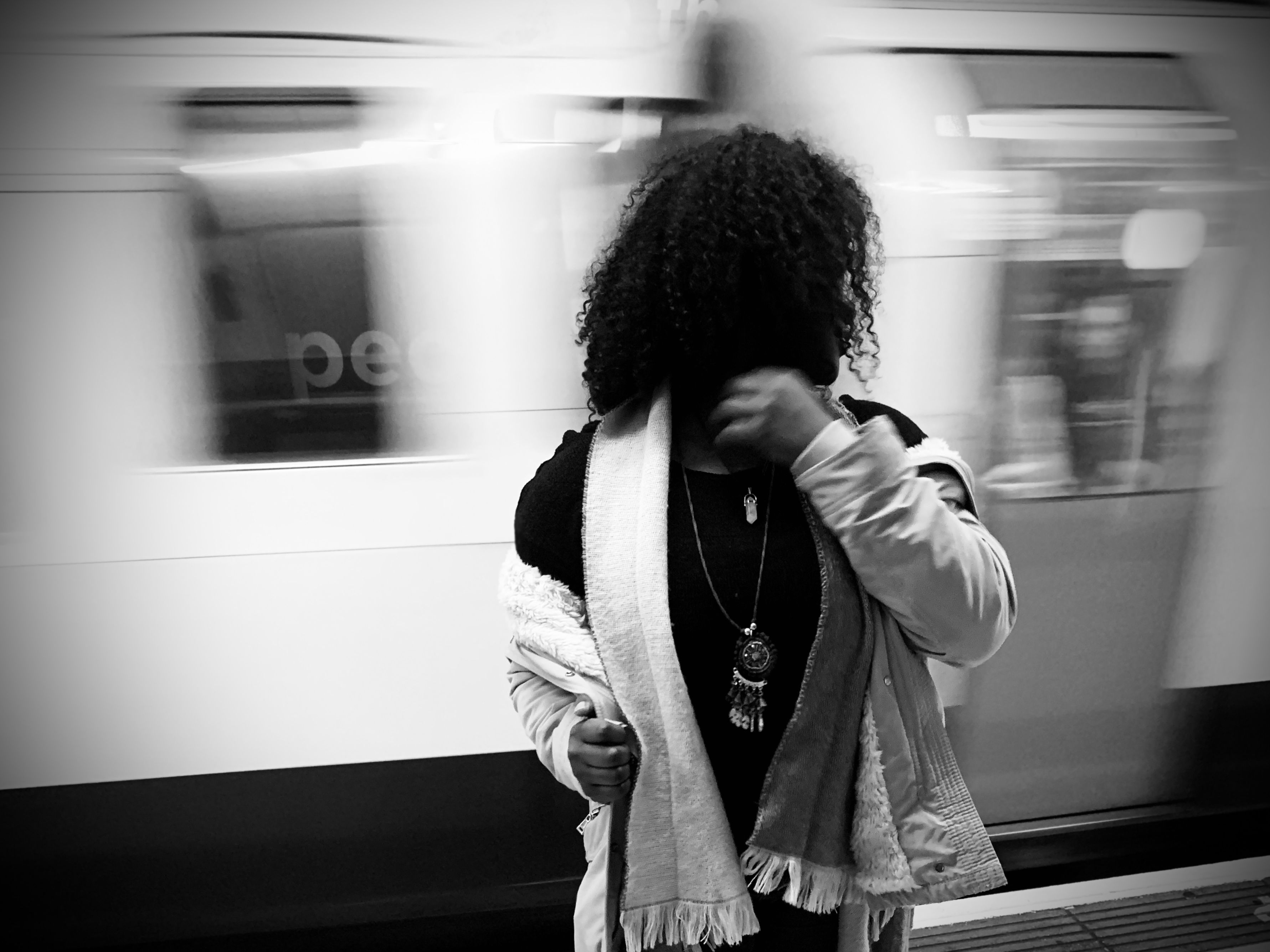 A woman with curly hair stands near a moving train on a London underground platform.