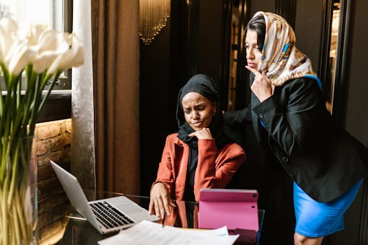 Two women in business attire discuss work at a laptop in a modern office.