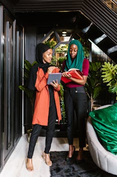 Two women in hijabs discussing and taking notes in modern office space.