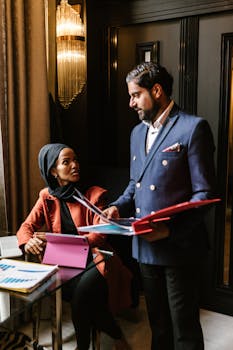 A man and woman in business attire discuss documents in an office setting.