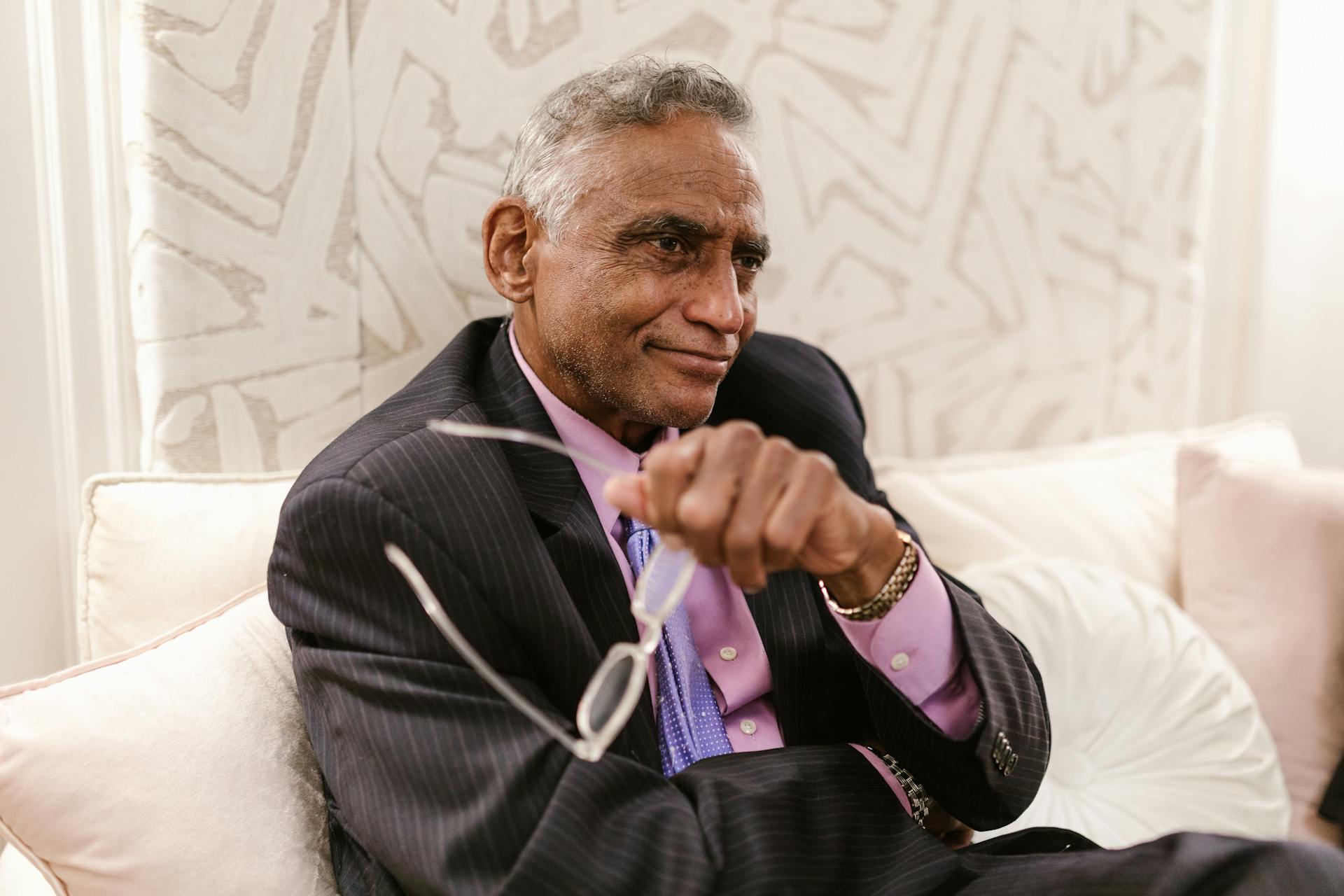 Senior man in business attire sitting comfortably indoors, holding eyeglasses with a relaxed smile.