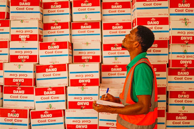 Man Working In A Warehouse Walking And Looking At Stack Of Boxes With A Notepad 