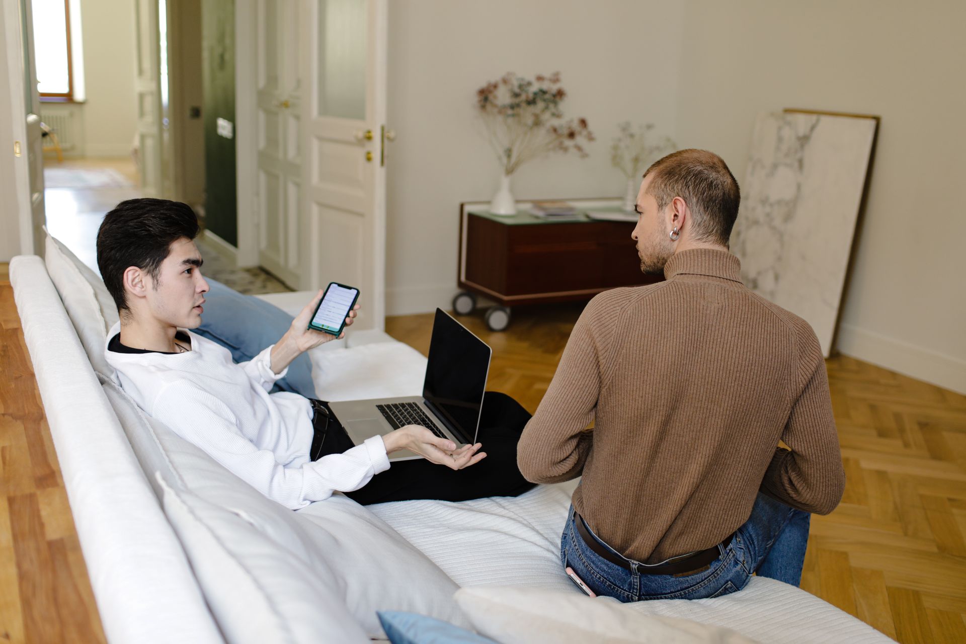 Two men engaging in conversation at home using laptop and phone, capturing modern communication.